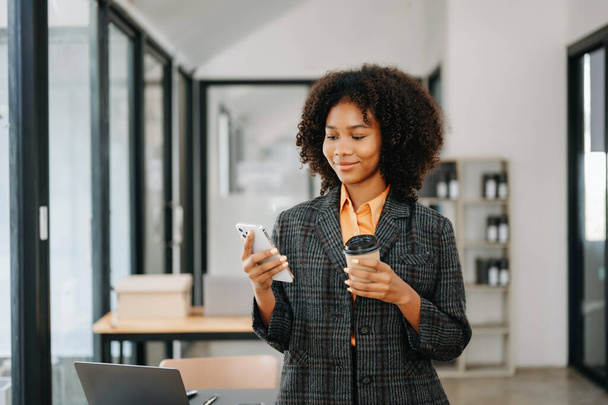 Confident African woman with a smile holding a phone and coffee at the modern office - Foto, Imagen