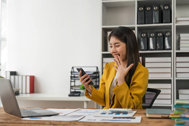 Ενθουσιασμένη επιχειρηματίας κερδίζει μετά την επίτευξη ανάγνωση ενός έξυπνου τηλεφώνου κάθεται σε ένα γραφείο στο γραφείο. - Φωτογραφία, εικόνα
