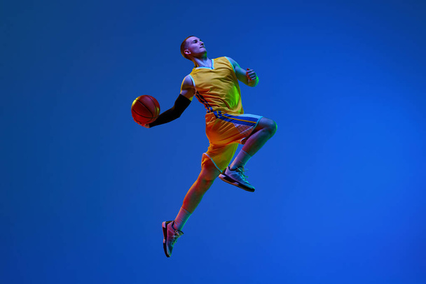 Νεαρός, μπασκετμπολίστας με κίτρινη στολή, ρίχνει μπάλα σε ένα άλμα σε μπλε φόντο στούντιο σε νέον φως. Έννοια του επαγγελματικού αθλητισμού, χόμπι, υγιεινό τρόπο ζωής, δράση, κίνηση - Φωτογραφία, εικόνα
