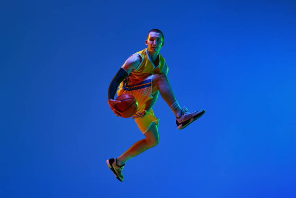 Dynamic image of professional male basketball player in motion, jumping with ball against blue studio background in neon. Winning goal. Concept of professional sport, hobby, healthy lifestyle, action - Фото, изображение