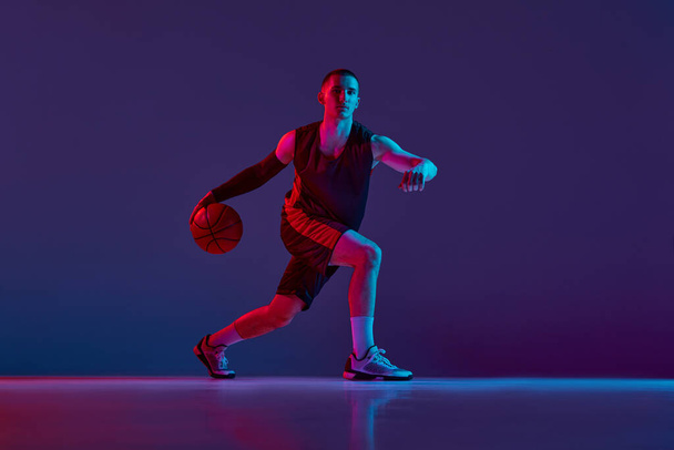 Joven deportista, jugador de baloncesto en uniforme durante el juego, driblando pelota contra fondo púrpura estudio en luz de neón. Concepto de deporte profesional, hobby, estilo de vida saludable, acción y movimiento - Foto, imagen