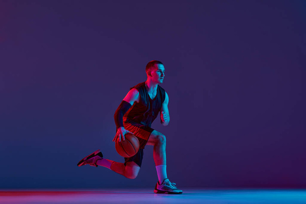 Concentrated young man, basketball player during game, training, dribbling ball against purple studio background in neon light. Concept of professional sport, hobby, healthy lifestyle, action, motion - 写真・画像