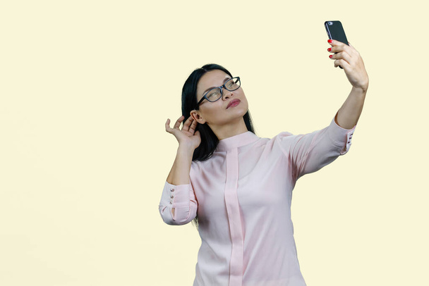 Cute young asian woman in glasses is taking a selfie on her smartphone. Isolated on yellow. - Фото, изображение