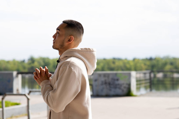 A young man in a white sweatshirt stands outdoors and stretches out his arms. Side view of an Asian man who stands against the background of the embankment. High quality photo - Photo, Image