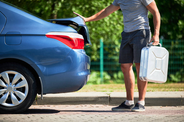 Człowiek wyjmujący kanister benzyną z bagażnika. Widok dorosłego mężczyzny wyjmującego cysternę z paliwem do zatankowania niebieskiego samochodu. - Zdjęcie, obraz