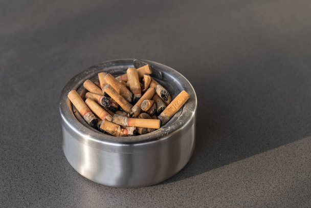 Schmutzige metallische Aschenbecher mit gebrauchten Zigarettenstummeln stapeln sich auf dem grauen Granitsteintisch. Viele Stups als Detail der Nikotinabhängigkeit. Konzept Raucherentwöhnung. - Foto, Bild