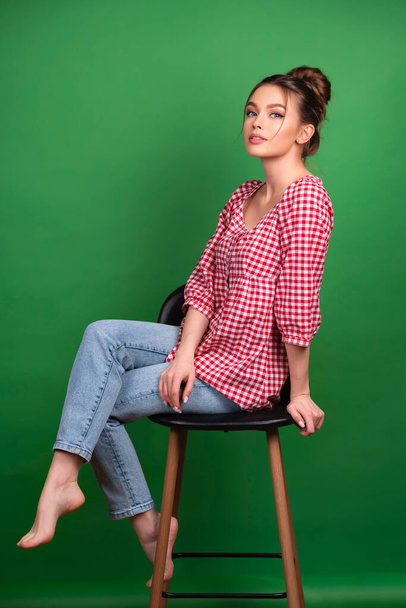 Hezká mladá dívka v kostkované košili a džíny pózování na židli na zeleném pozadí izolované.Studio práce s atraktivní model. - Fotografie, Obrázek
