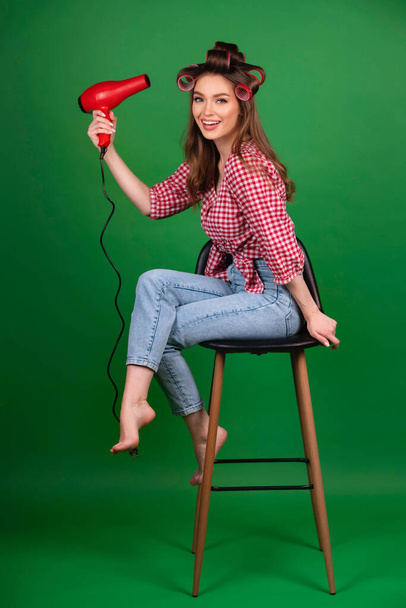 Ładna młoda uśmiechnięta dziewczyna w dużych czerwonych lokówkach susząca włosy z czerwoną suszarką. Studio pracy z atrakcyjnym modelem w kratkę czerwonej koszuli i dżinsy pozowanie na zielonym tle izolowane. - Zdjęcie, obraz