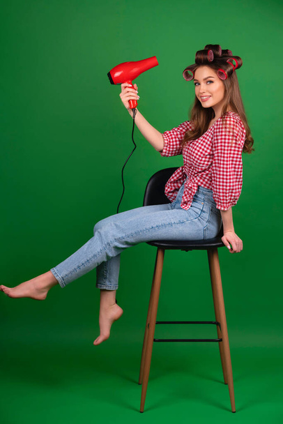 Όμορφη νεαρή χαμογελαστή κοπέλα με μεγάλα κόκκινα μπικουτί να στεγνώνουν τα μαλλιά με κόκκινο στεγνωτήρα. Studio εργασία με ελκυστικό μοντέλο σε καρό κόκκινο πουκάμισο και τζιν ποζάρουν σε πράσινο φόντο απομονωμένο. - Φωτογραφία, εικόνα