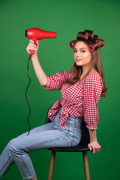 Pretty young smiling girl in big red curlers drying hair with red dryer. Studio work with attractive model in checkered red shirt and jeans posing  on green background isolated. - Фото, зображення