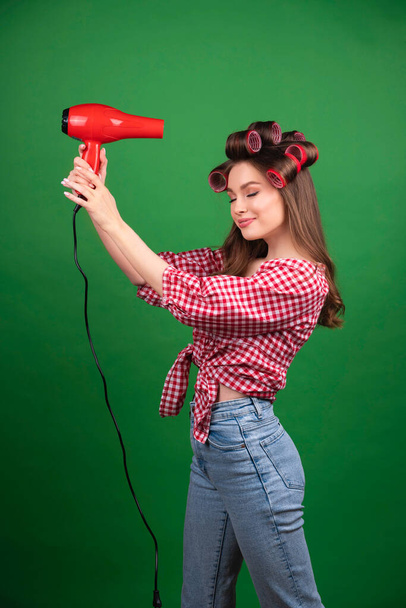 Pretty young smiling girl in big red curlers drying hair with red dryer. Studio work with attractive model in checkered red shirt and jeans posing  on green background isolated. - Photo, Image
