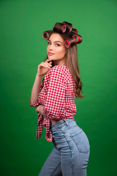 Pretty young smiling girl in big red curlers drying hair with red dryer. Studio work with attractive model in checkered red shirt and jeans posing  on green background isolated. - Фото, изображение