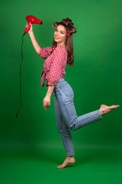 Vrij jong lachend meisje in grote rode krulspelden die haar drogen met rode droger. Studio werk met aantrekkelijk model in geruit rood shirt en jeans poseren op groene achtergrond geïsoleerd. - Foto, afbeelding
