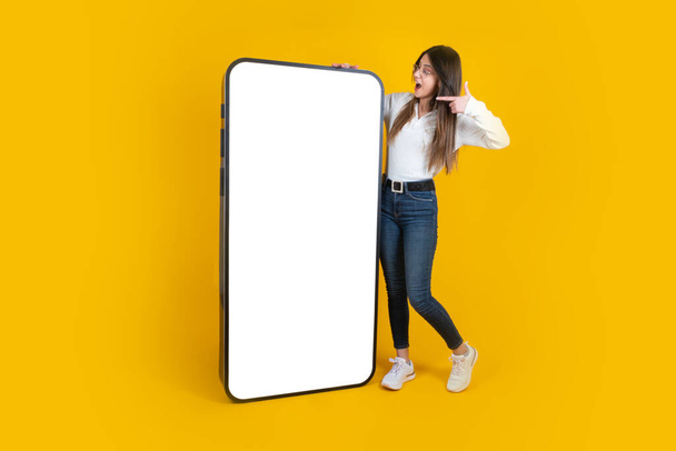 Woman pointing big huge blank screen mobile phone. Full body length amazed young lady standing over isolated yellow studio background. Wearing jeans and white shirt. Smartphone mockup, copy space. - Foto, Bild