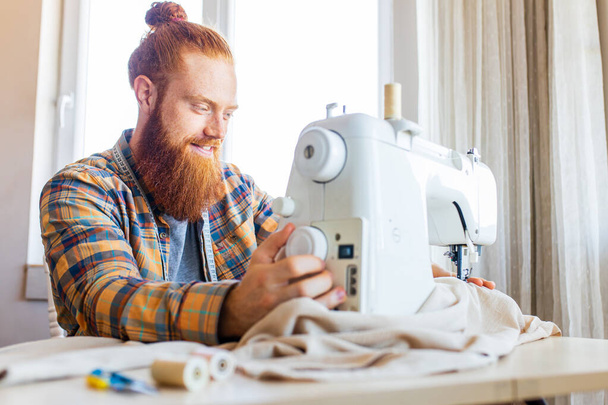 handsome redhaired man with long beard sews at a sewing machine at home studio. - Фото, зображення
