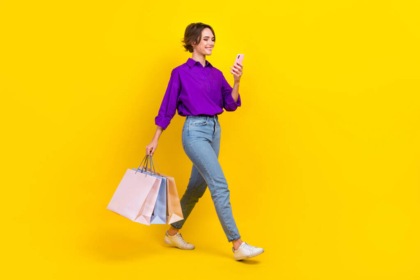 Full length photo of pretty sweet lady wear violet shirt walking holding bargains texting gadget isolated yellow color background. - Foto, imagen