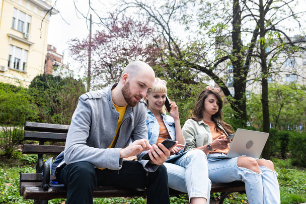 Three students on a bench in the park study talking typing on laptop notebook and writing. Education concept. Young colleagues working on ideas and solutions for new strategy business - Fotografie, Obrázek