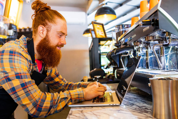 Professional barista young redhaired ginger bearded man in black apron working in coffee shop. - Fotó, kép