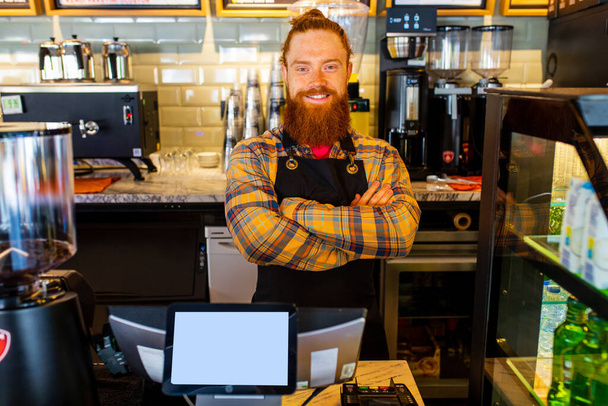 Professional barista young redhaired ginger bearded man in black apron working in coffee shop. - 写真・画像