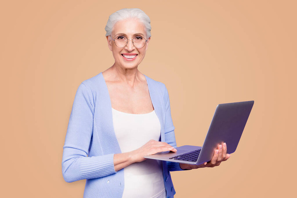Gris pelo viejo agradable hermosa mujer sonriente alegre con gafas que sostienen portátil aprendizaje curso en línea. Aislado sobre fondo violeta púrpura
. - Foto, Imagen