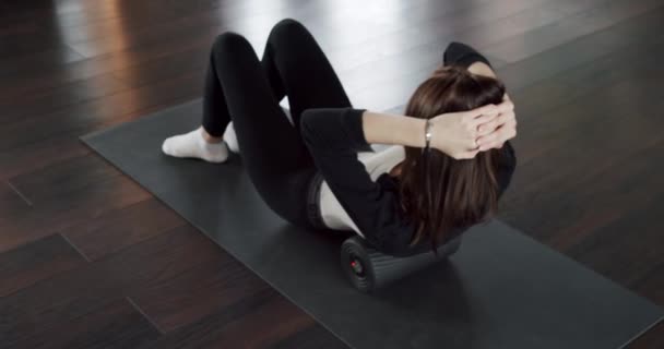Tracking shot of female stretching legs on foam roller while exercising on mat during training in light studio. Sportswoman doing crunches with sport equipment. Girl warming up on the fitness mat. - Footage, Video