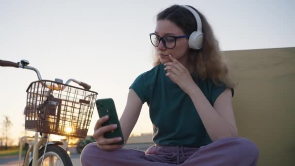 Millennial meisje zit op een bankje in het park met een telefoon in haar handen, maakt een sefi en luistert naar muziek in draadloze koptelefoon bij zonsondergang. Fietsen door het stadspark op een warme zomeravond. - Video