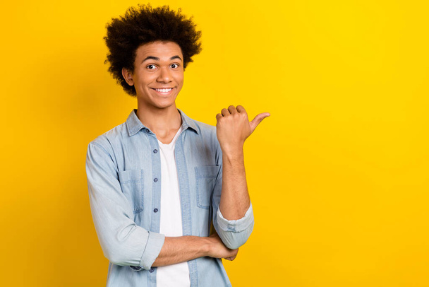 Photo of cool cheerful guy dressed denim shirt pointing thumb empty space isolated yellow color background. - Foto, afbeelding