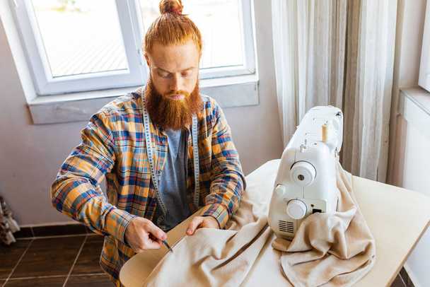 handsome redhaired man with long beard sews at a sewing machine at home studio. - Фото, зображення