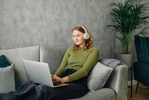 Headphones, laptop and woman on a home sofa listening to music or audio while streaming online. Calm female person relax on couch to listen to radio or song with internet connection and technology - 写真・画像