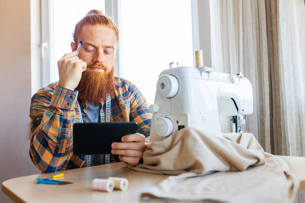 handsome redhaired man with long beard sews at a sewing machine at home studio. - Zdjęcie, obraz