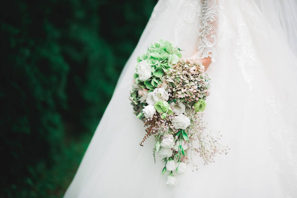 Beautiful bride in elegant white dress holding bouquet posing in park. - Fotoğraf, Görsel