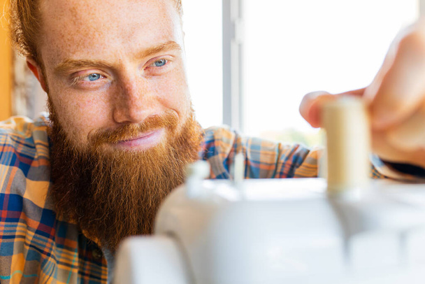 handsome redhaired man with long beard sews at a sewing machine at home studio. - Fotografie, Obrázek