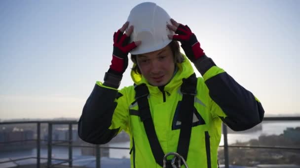 Portrait of an industrial climber builder man in bright overalls and gear puts on a working white hard hat while standing on the roof at sunset - Footage, Video