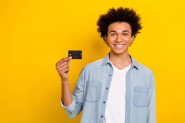 Photo of cheerful cute guy dressed denim shirt rising bank card empty space isolated yellow color background. - Φωτογραφία, εικόνα
