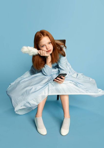 Portret met een verdrietige prinses, koningin met rood haar in feestelijke jurk met smartphone over een blauwe achtergrond. Concept van de moderne, technologieën, gadgets, middeleeuwse tijdperk, schoonheid, mode, emoties, reclame - Foto, afbeelding
