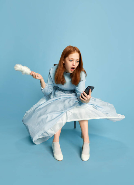 Princesa, reina de pelo rojo vistiendo vestido festivo sosteniendo smartphone con la cara sorprendida sobre fondo azul. Concepto de moderno, tecnologías, gadgets, época medieval, belleza, moda, emociones, ad - Foto, imagen