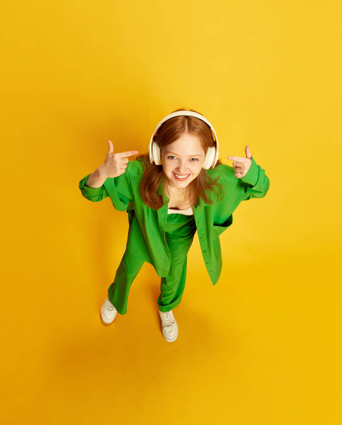 Happy girl, teenager with red hair wearing stylish green costume wearing wireless headphones over yellow studio background. Top view. Concept of youth, fashion, music, lifestyle, hobby, emotions, ad - Photo, Image