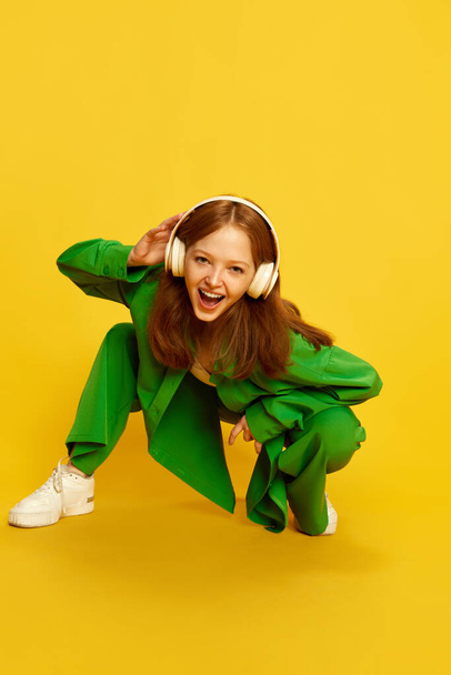 Funny girl, teenager with red hair wearing wireless headphones listening to music over yellow studio background. Top view. Concept of youth, fashion, lifestyle, hobby, human emotions, ad - Foto, imagen