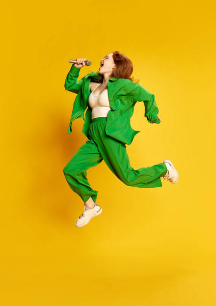 Delight, happiness. Emotional beautiful girl with red hair in wireless headphones jumping over yellow background. Music, hobby, happy positive emotions and active lifestyle concept - Photo, Image
