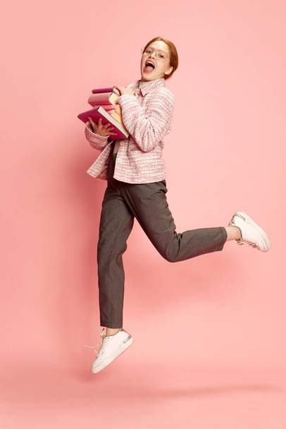 Jolie jolie étudiante aux cheveux roux portant l'uniforme, tenant des livres sautant et souriant sur fond de studio rose. Concept de vie étudiante, études, école, éducation à l'étranger, apprentissage, publicité - Photo, image