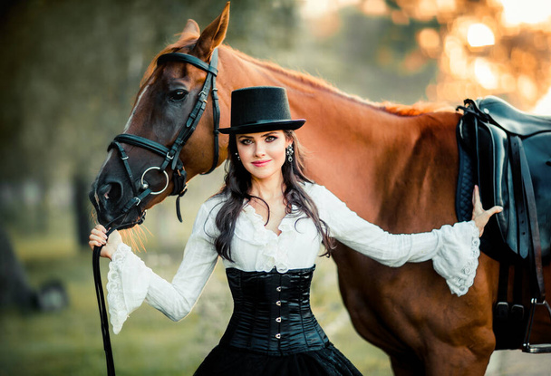 Portret van prachtige Fashion gothic meisje wandelen een paard .Fantasy kunstwerk en sprookje.Amazing brunette model in zwart-wit jurk en hoed poseren. - Foto, afbeelding