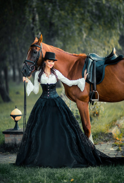 Retrato de magnífica chica gótica de moda caminando un caballo. Fantasía obra de arte y hada. Increíble modelo morena en vestido blanco y negro y sombrero posando. - Foto, Imagen