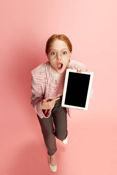 One young girl, student wearing eyeglasses holding a tablet and pointing at empty gadget screen over a pink background. Concept of education, online, student life, human emotions, social networks, ad - Foto, imagen