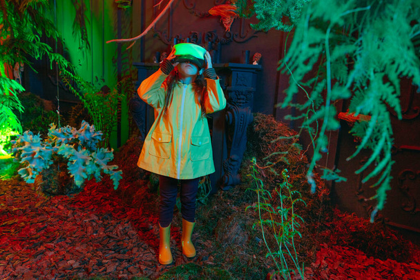 Κοριτσάκι με φωτεινά γυαλιά εικονικής πραγματικότητας που στέκεται στο δωμάτιο με φυτά και δέντρα με φώτα νέον σαν προσομοιωτής εικονικής πραγματικότητας. επιλεκτική εστίαση. - Φωτογραφία, εικόνα