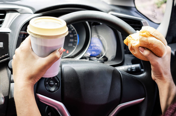 verboten und gefährlich mit Nahaufnahme der Hand einer Frau, die Burger und Kaffee in der Hand hält, rücksichtslos isst und trinkt, während sie Auto fährt - Foto, Bild