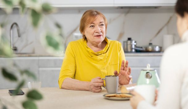 Indignant elderly woman with cup of tea sits in kitchen and listens to daughter.Women talk about family difficulties.Concept of troubled family relationships - Foto, Bild