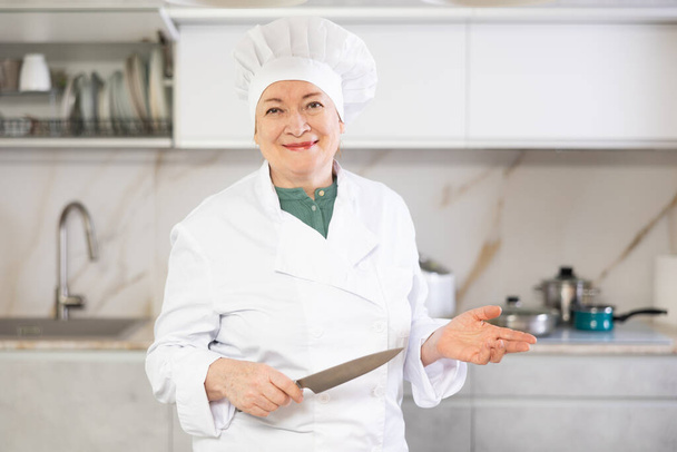 In der geräumigen und hellen Küche hält eine ältere Küchenchefin ein Messer in der Hand und wartet darauf, dass Gäste einen Kochkurs für Kinder leiten. - Foto, Bild
