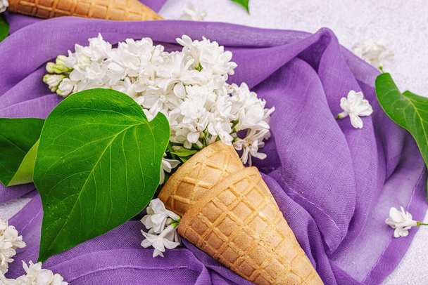 Белые лиловые цветы в вафельных рожках мороженого на светлом каменном бетонном фоне. Плоская кровать, фиолетовая салфетка, традиционная весенняя концепция, крупным планом - Фото, изображение