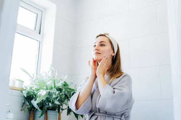 Young woman in bathrobe looking in mirror and doing manual self massage of face in the bathroom. Natural procedures for skin care. Home Beauty treatment, face gymnastics for youth. Selective focus - Photo, image