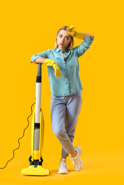 Mujer joven y cansada con aspiradora sobre fondo amarillo - Foto, imagen